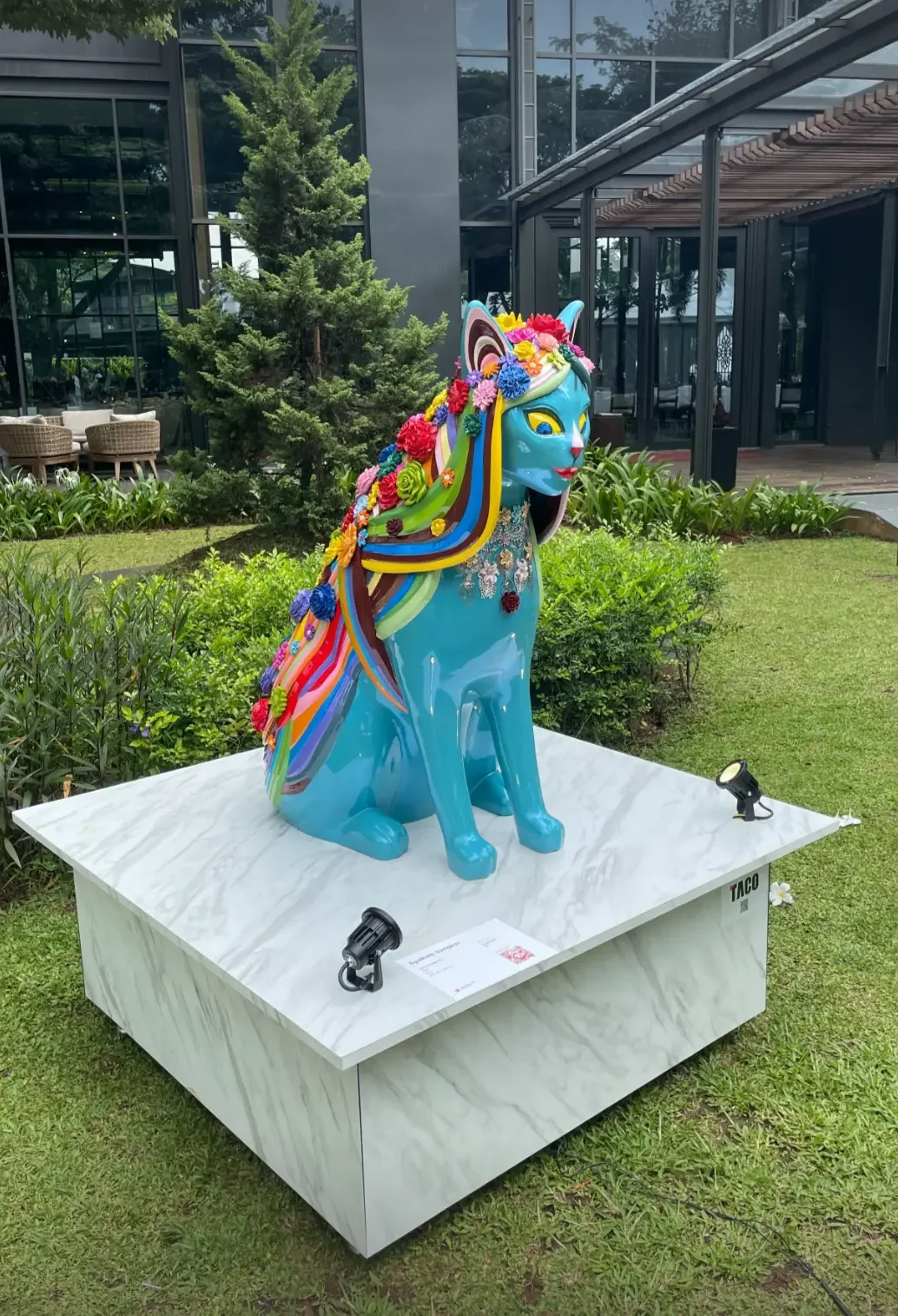 A large glossy turquoise cat sculpture adorned with colourful flowers and rainbow-striped wings, displayed on a marble plinth in a garden setting with the Plataran restaurant building in the background