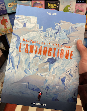 Close-up of a hand holding a graphic novel titled “Des ronds de serviette pour l’Antarctique” featuring an illustrated icy Antarctic landscape on the cover.