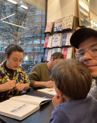 Inside a bookshop, an illustrator signs books at a table while a child and adult sit nearby. Bookshelves and large windows are visible in the background.