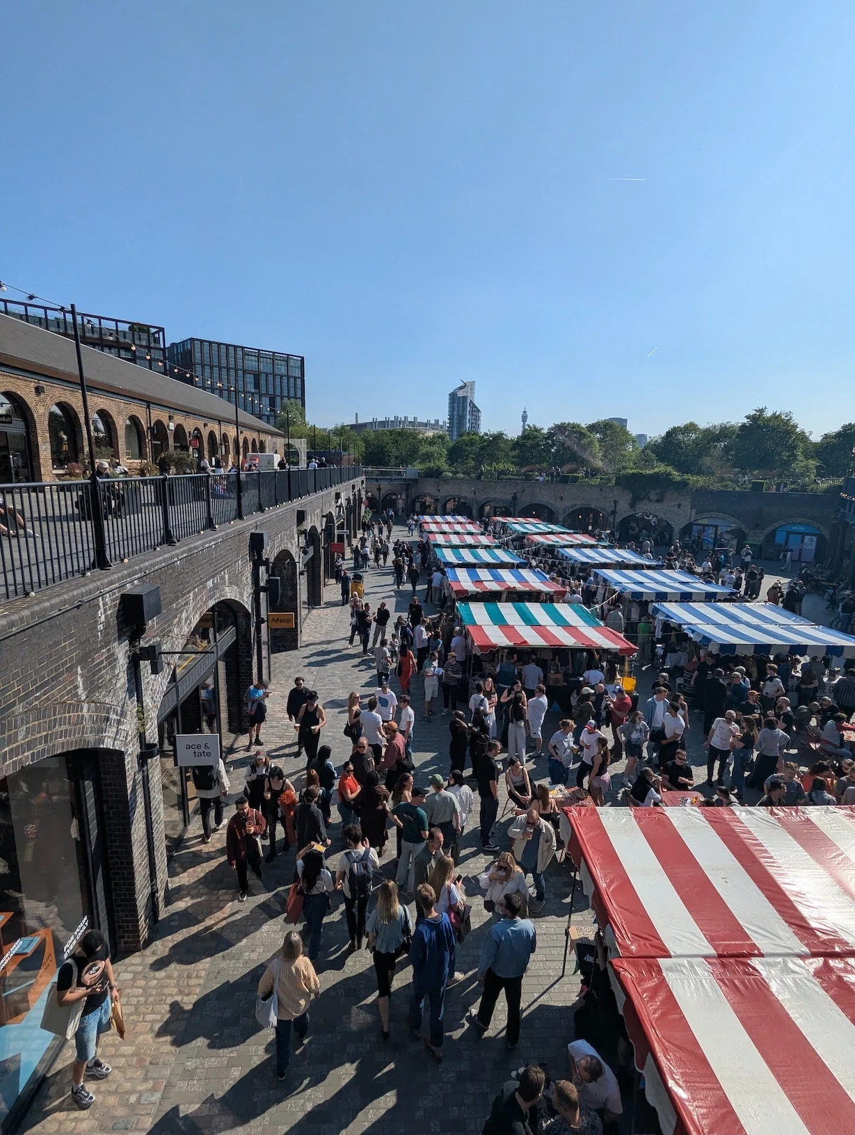 Aerial view of the Independent Label Market at Coal Drops Yard, Kings Cross, London, showing colourful striped market stalls and a large crowd on a sunny day, with the Victorian brick arches of Coal Drops Yard visible