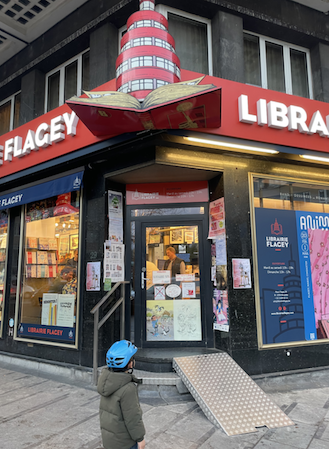 Exterior of Librairie Flagey bookshop in Brussels with a red awning and signage. A child wearing a blue helmet stands outside near the entrance, looking towards the shop.