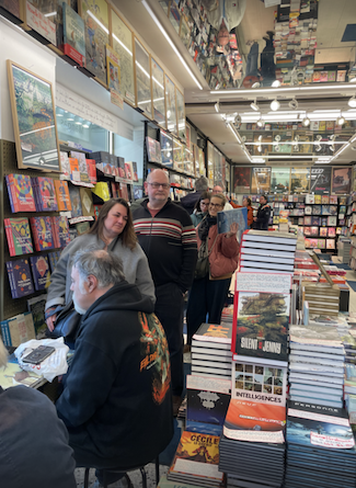 Interior of a busy bookshop with people standing in line among tables stacked with books. Shelves filled with titles line the walls.
