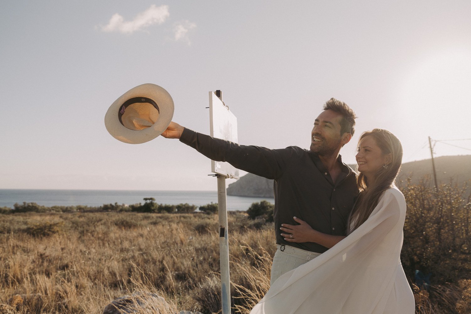 A couple stands outdoors on a grassy landscape near the coast, smiling and embracing each other, with the man holding a sunhat in the air, during sunset or late afternoon.