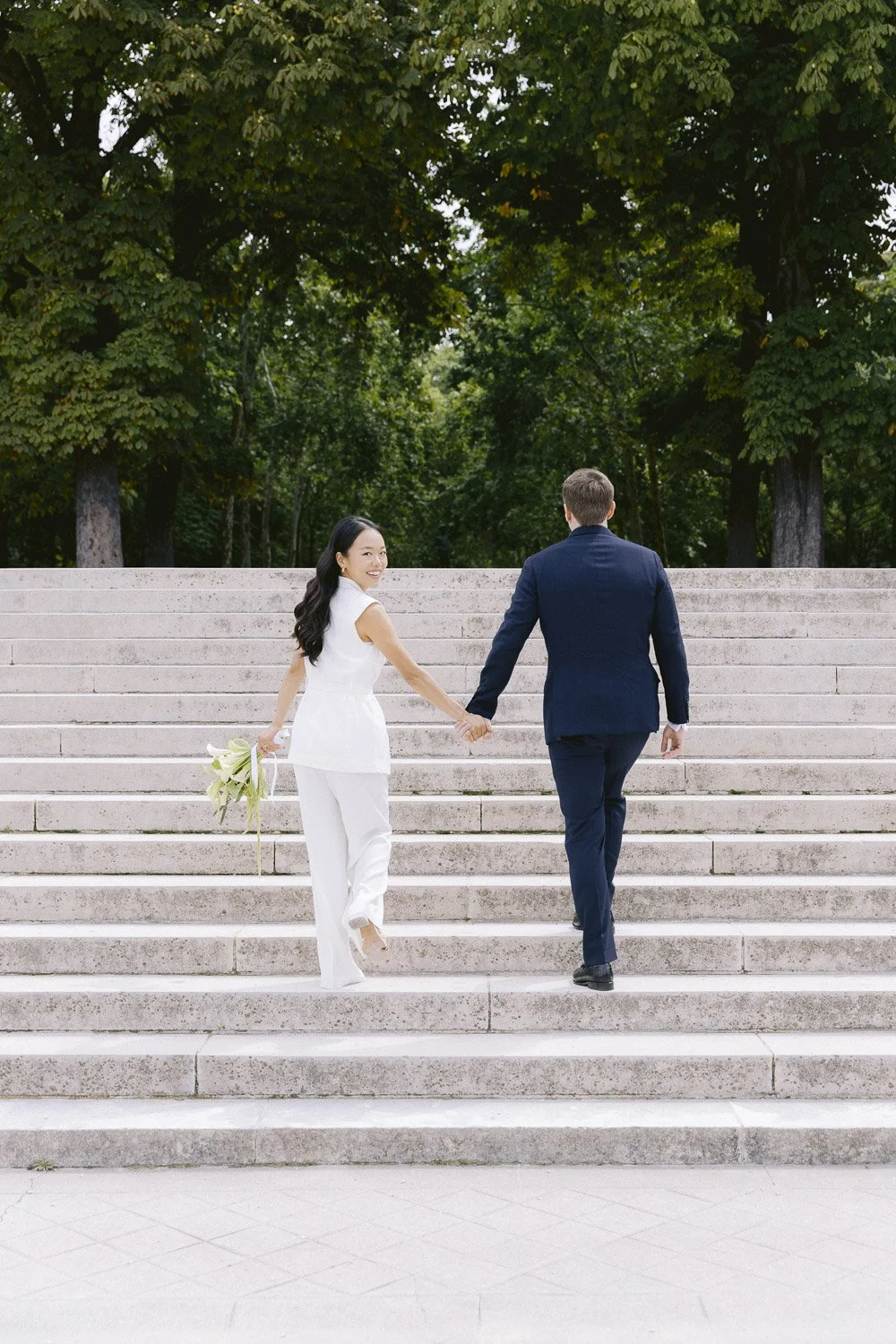 A couple holding hands and walking up stairs outdoors, with the woman smiling and holding a bouquet, and trees in the background.