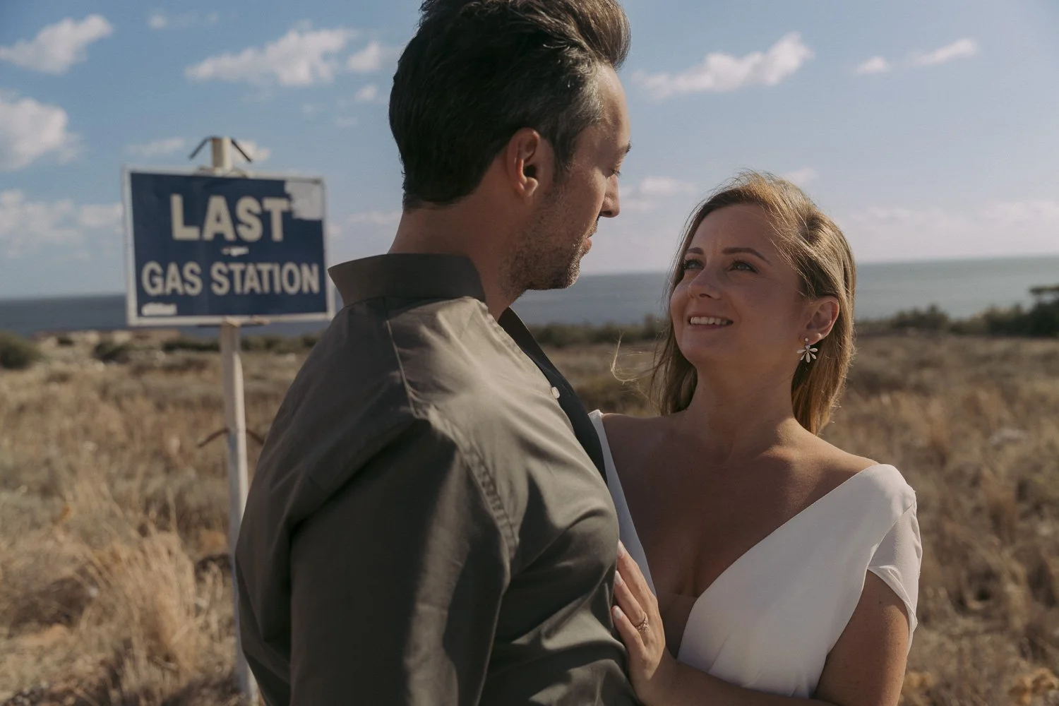 A couple standing close together outdoors near the coast, with the man looking down at the woman and the woman smiling at him, in front of a sign that reads "Last Gas Station."