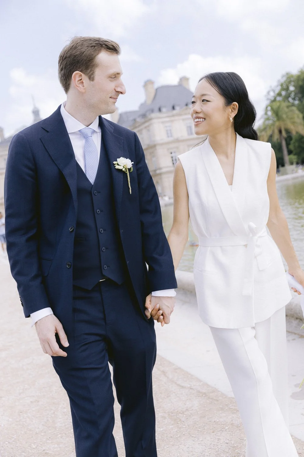 A couple holding hands, smiling, outdoors near a historic building, dressed in formal attire, with the man in a navy suit and the woman in a white outfit.