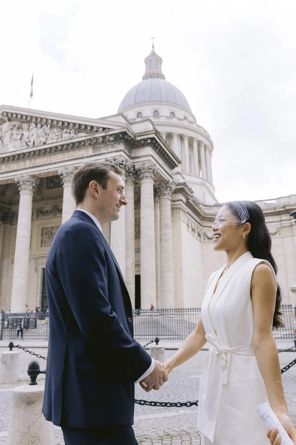 A couple in wedding attire holding hands and smiling at each other outside a historic building with classical columns and a dome.