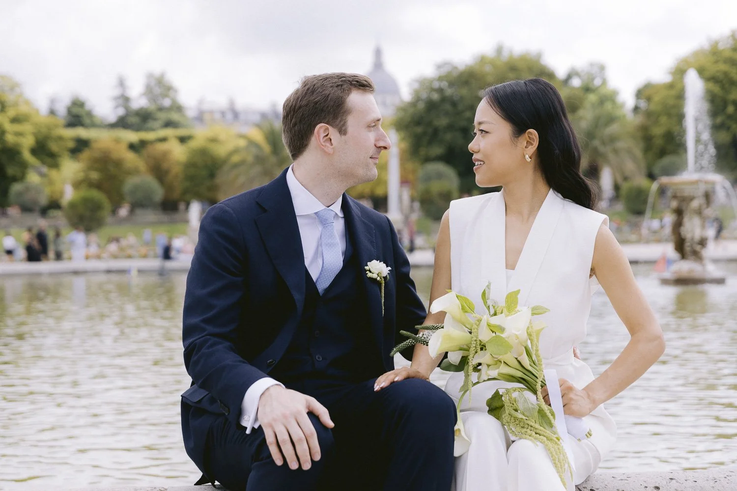 A newlywed couple sitting by a fountain in a park, looking into each other's eyes, with trees and a distant cityscape in the background.