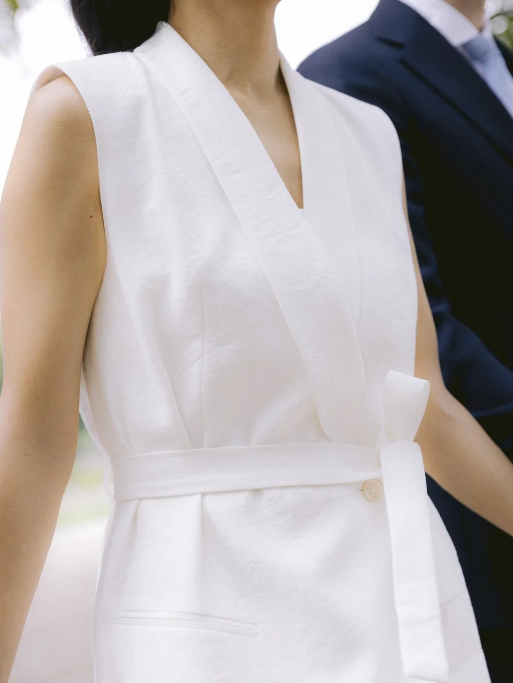 Close-up of a woman wearing a white sleeveless dress with a V-neckline, standing next to a man in a dark suit. The background is blurred with natural outdoor lighting.