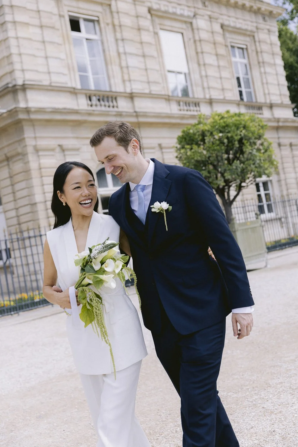 A newlywed couple, a woman in a white dress and a man in a navy suit, smiling and walking together outside a historic building with large windows and a tree in the background.