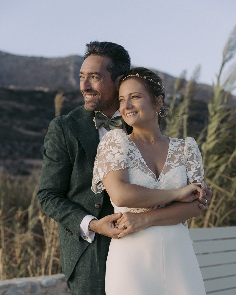 A bride and groom are outdoors, smiling and embracing each other. The groom is wearing a dark suit with a bow tie, and the bride is in a white wedding dress with lace sleeves and a headband. Background includes hills and tall grasses.