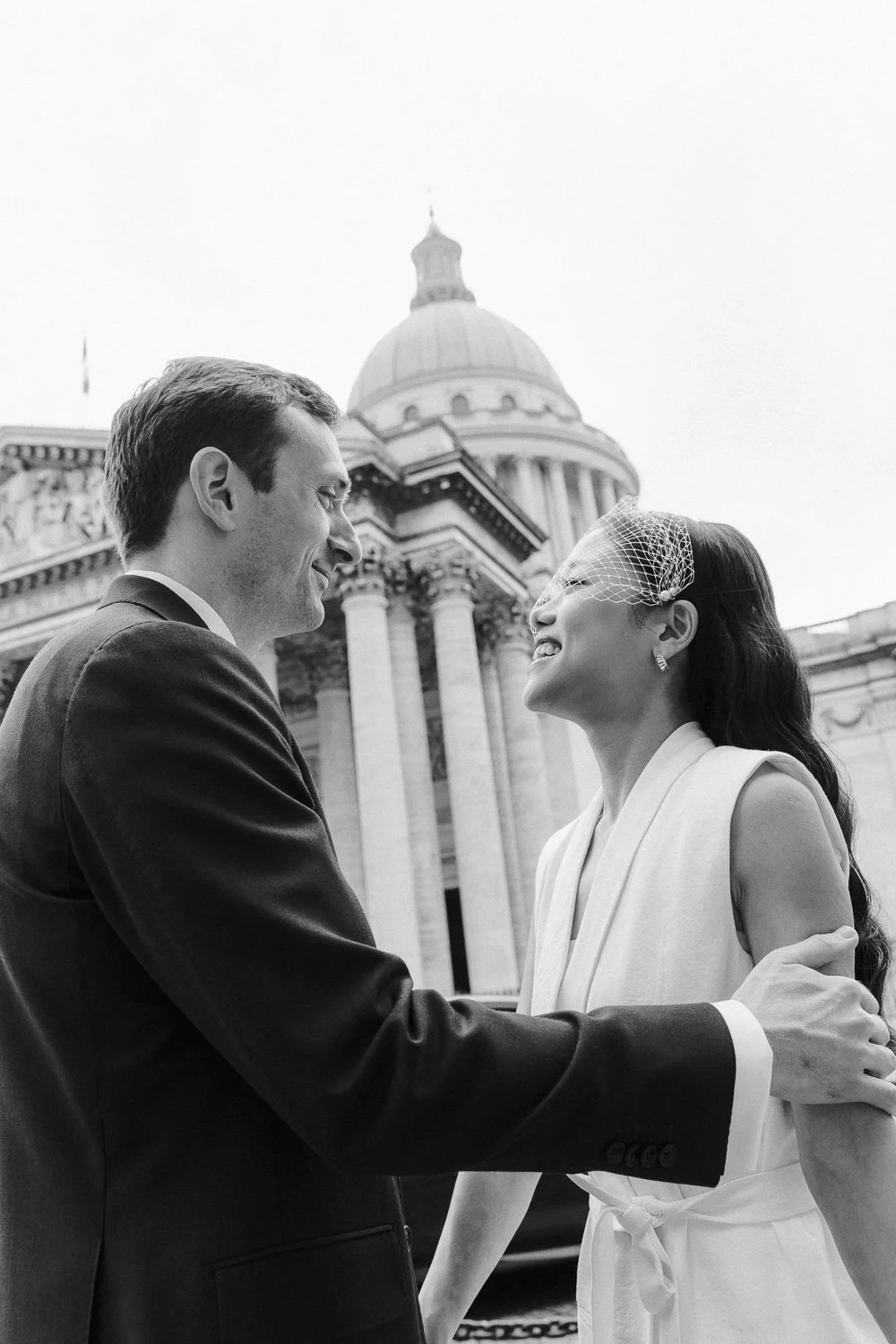 A couple, a man and woman, smiling and looking at each other, standing outdoors in front of a government building with a domed roof and columns. The woman is wearing a dress with a small veil, and the man is in a suit.