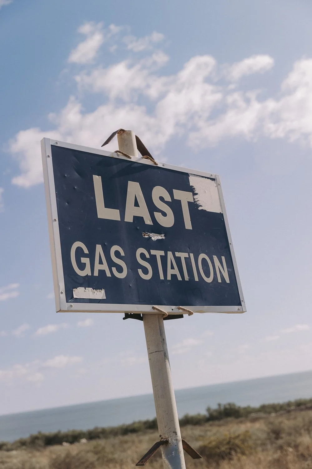 A blue sign that reads "Last Gas Station" with some wear and tear, mounted on a silver pole against a partly cloudy sky and ocean in the background.