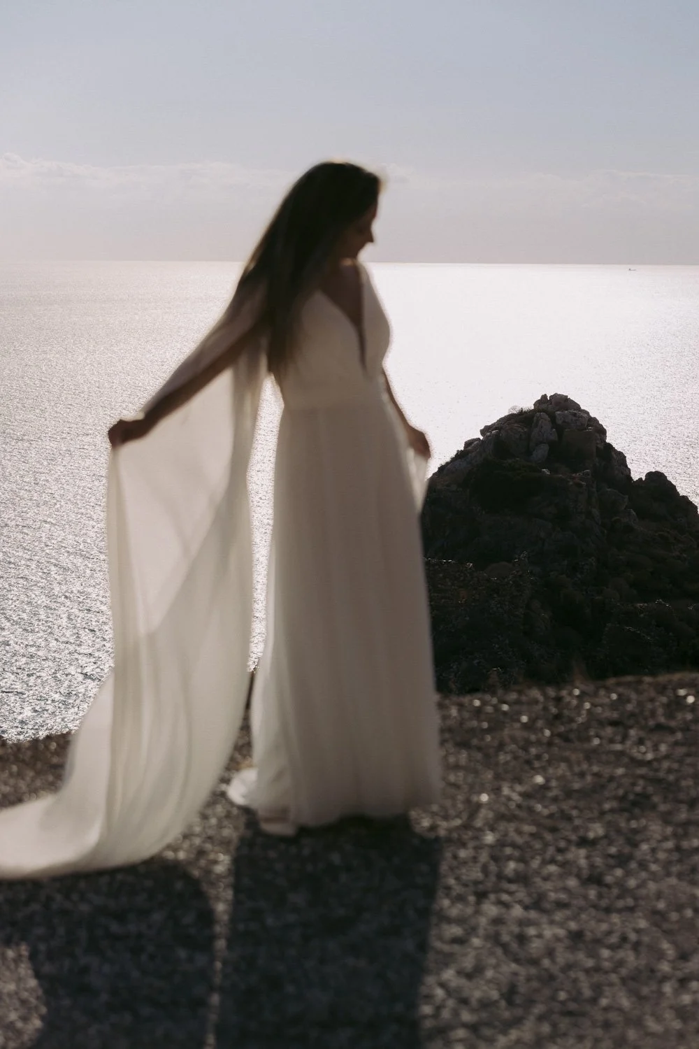 A woman in a long white dress standing on a rocky shoreline by the water, with the sun reflecting off the water, creating a bright background.
