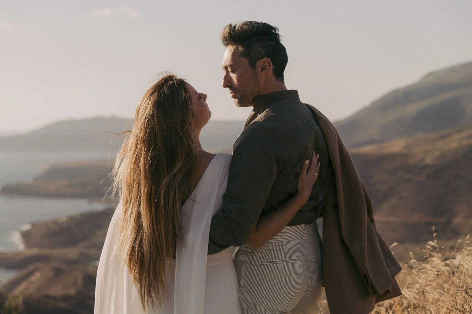 A man and woman standing close together outdoors near water and hills, gazing into each other's eyes during what appears to be sunset.