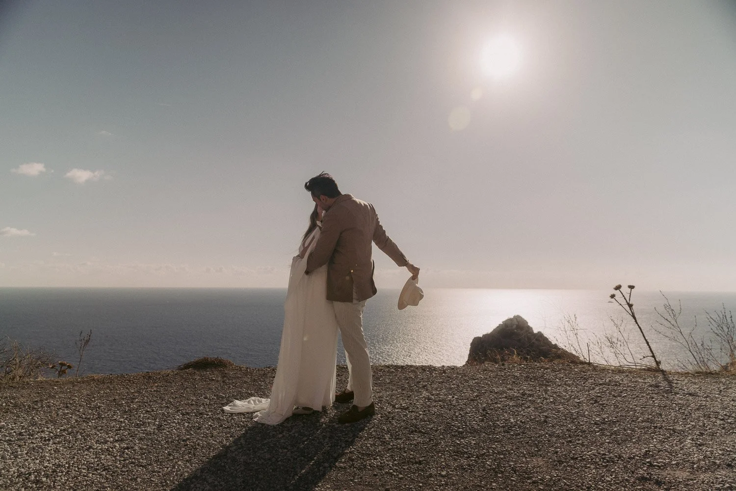 A couple in wedding attire embracing on a cliff with the ocean and a bright sky in the background.