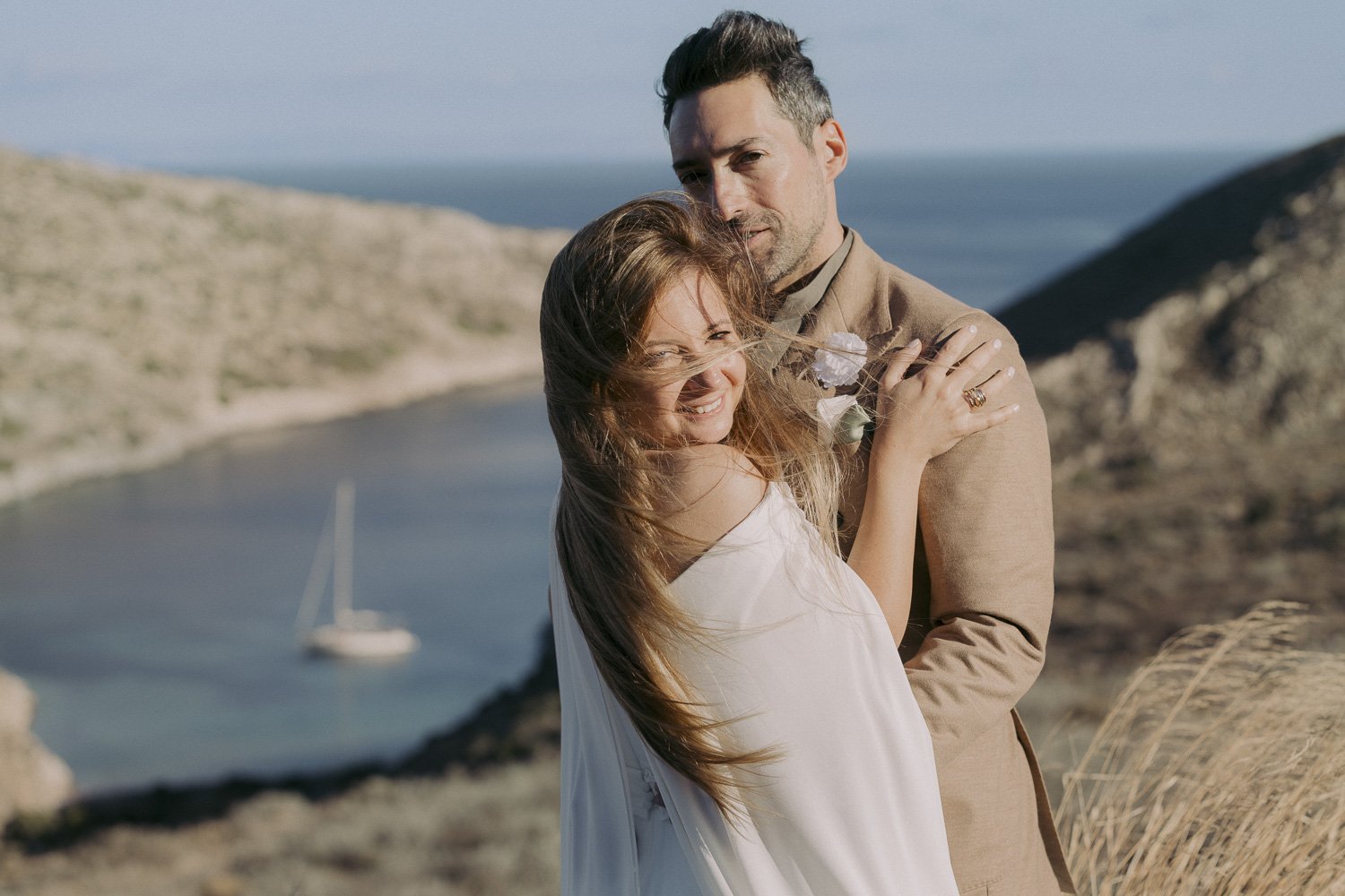 A couple embracing outdoors near a body of water with a sailboat, hills, and the sea in the background.