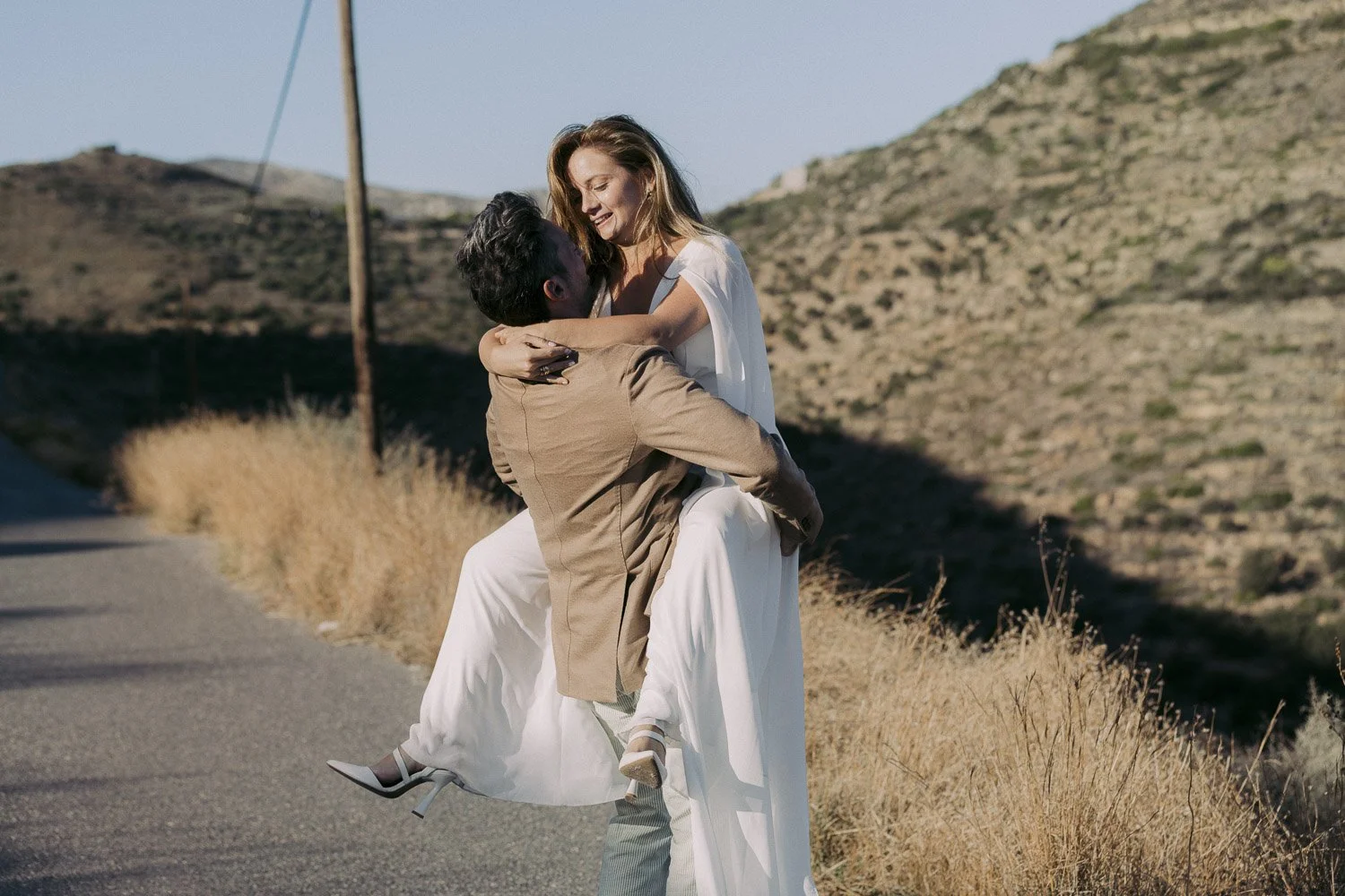 A man lifts a woman in his arms on a rural road with hills in the background. They are smiling and appear happy.