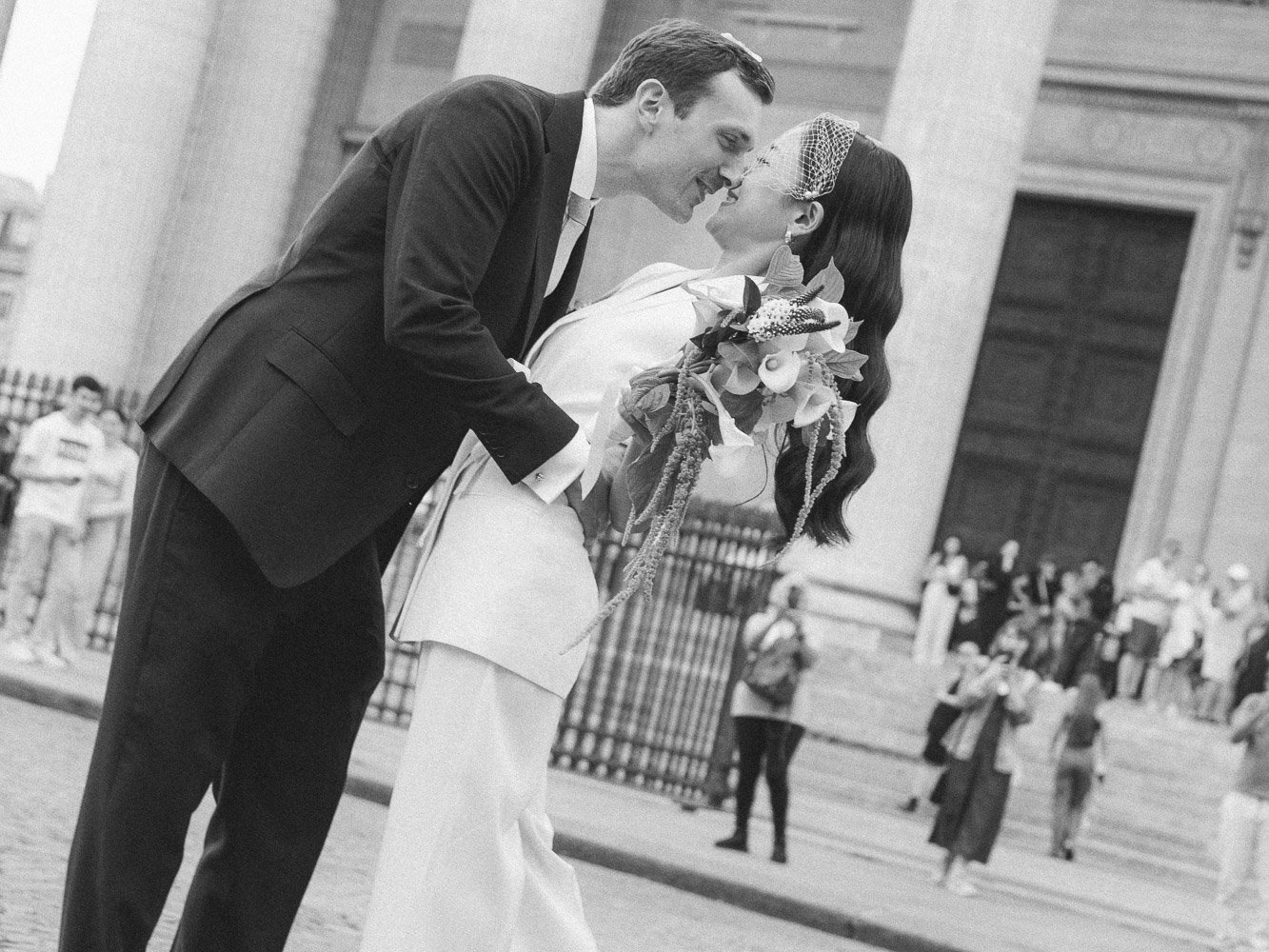 A groom and bride sharing a kiss on their wedding day outside a large building with stairs, with several people in the background.