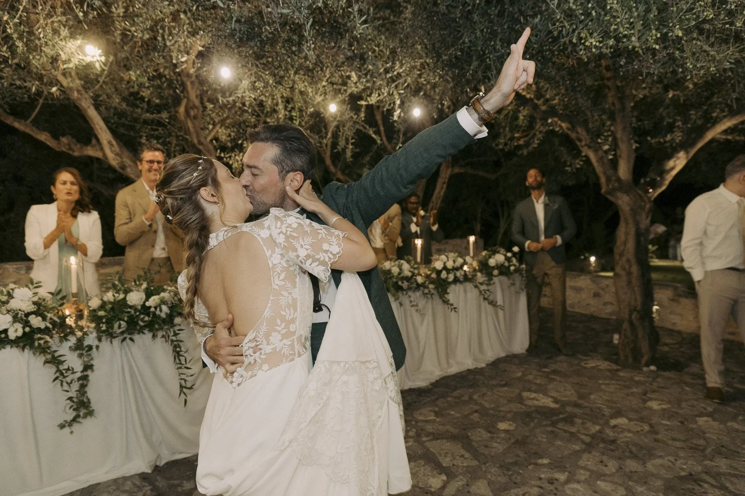 A bride and groom kiss during their outdoor wedding reception at night, surrounded by friends and family under string lights and trees.