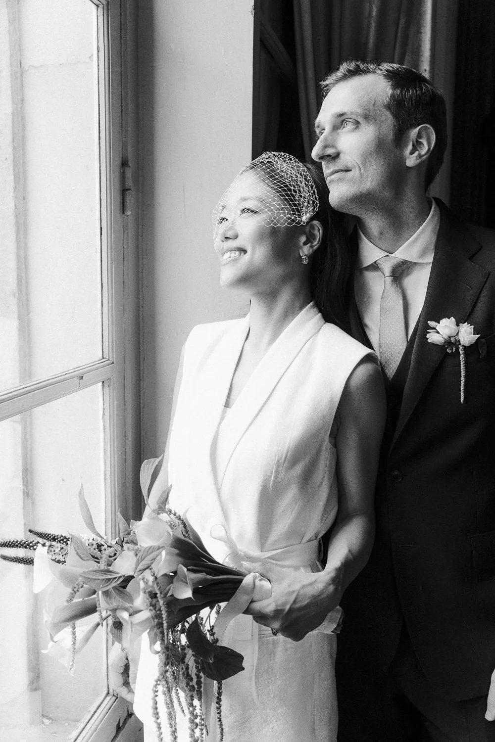 A bride and groom looking out a window, smiling. The bride holds a bouquet of flowers, and the groom wears a boutonniere. The scene is in black and white.