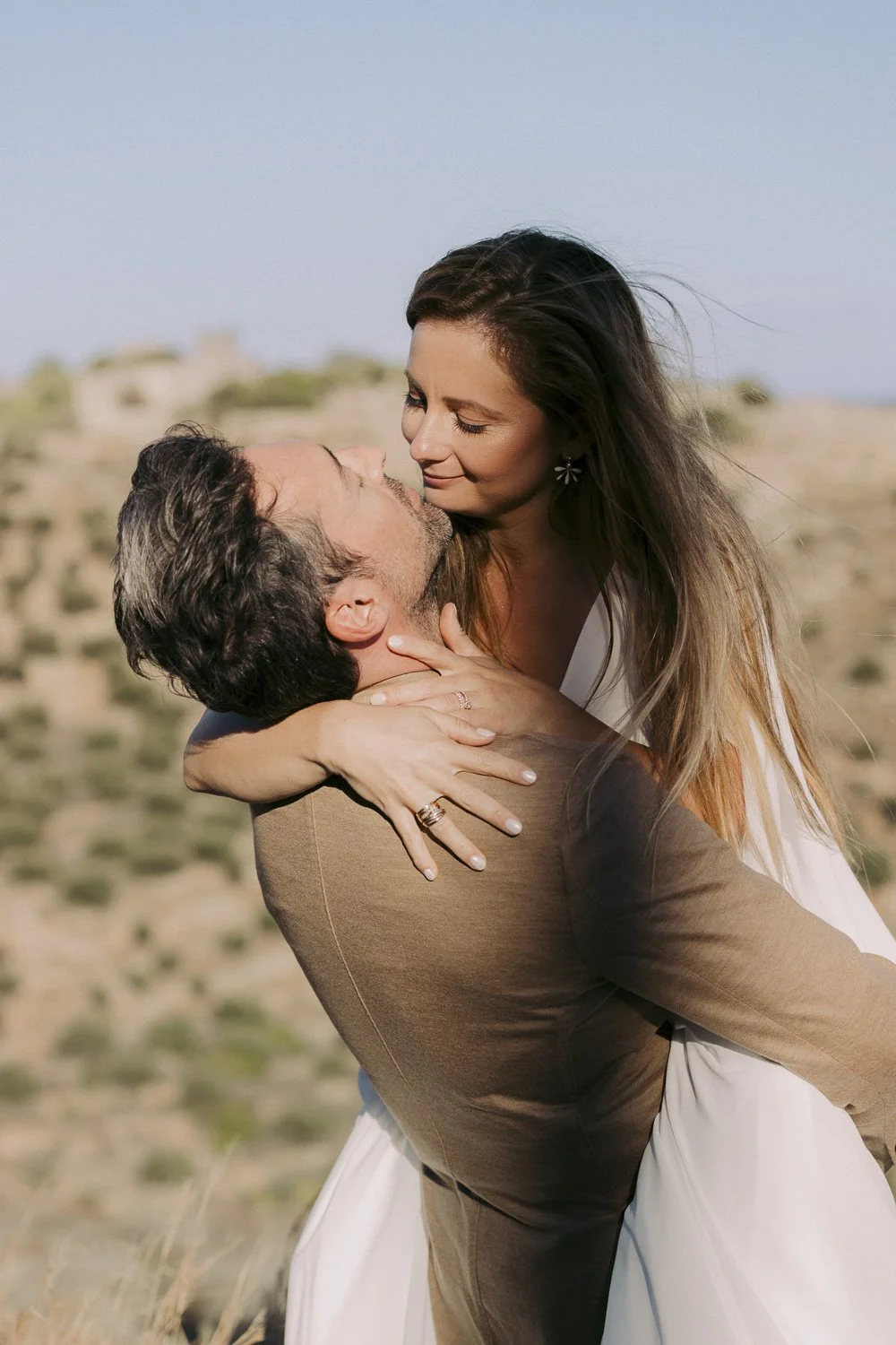 A man and woman sharing a romantic embrace outdoors, with desert-like terrain in the background, during daytime.