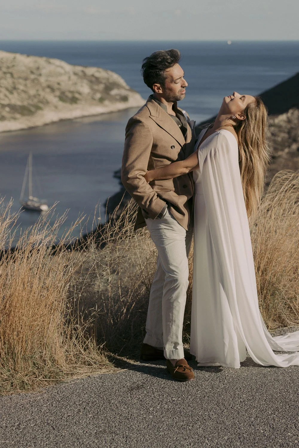 A couple embracing on a grassy cliff overlooking the ocean with a sailboat in the water and rocky cliffs in the background, during daytime.