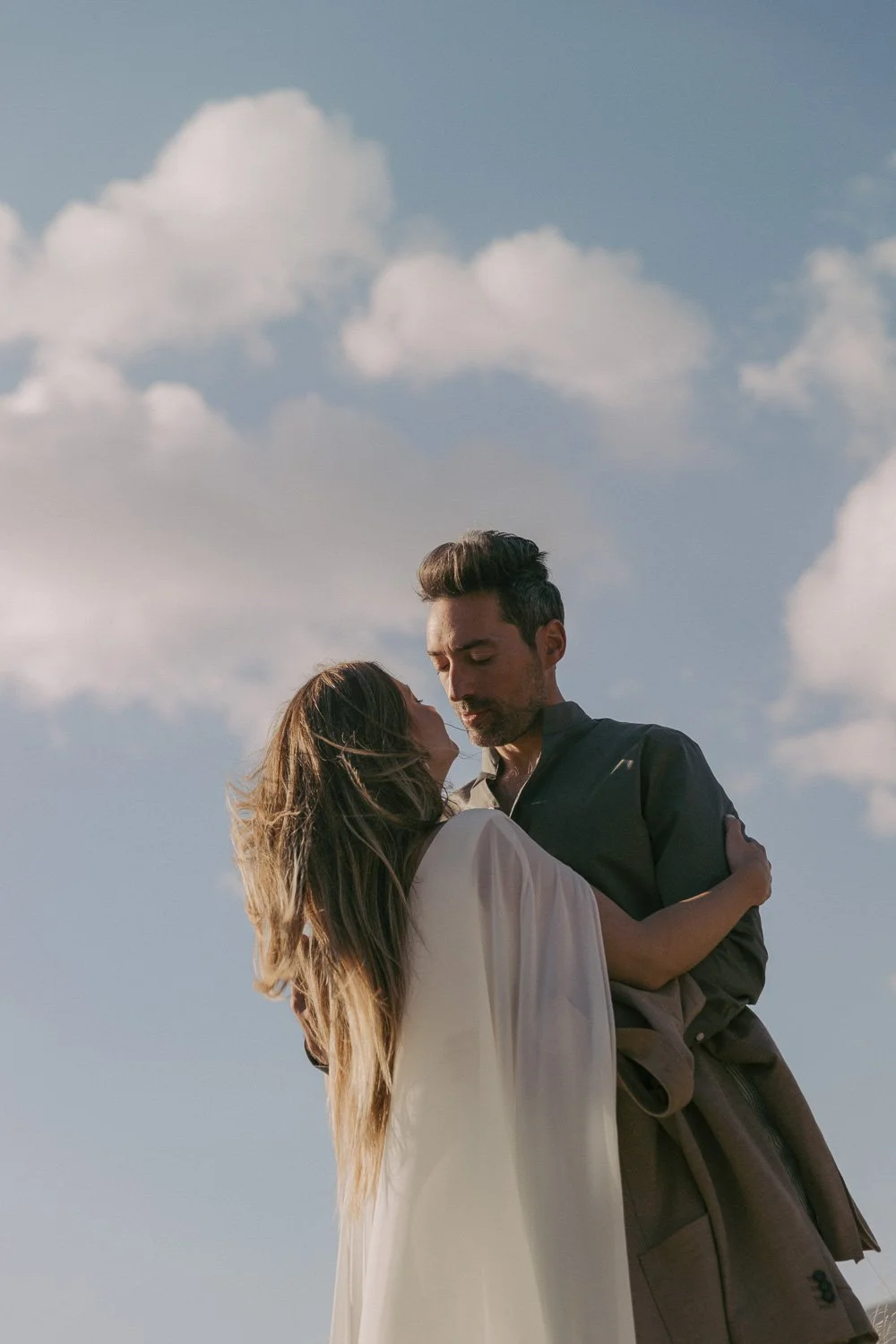 A couple sharing a romantic moment outdoors beneath partly cloudy sky, with the woman holding onto the man's shoulders and the man gently holding her waist.