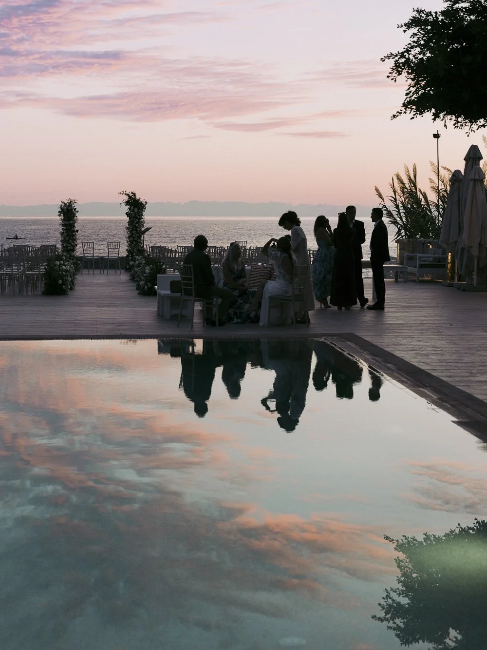 A group of people gathered on a deck overlooking the water during sunset, with the sky filled with pink and purple clouds, and their reflections visible in the pool below.
