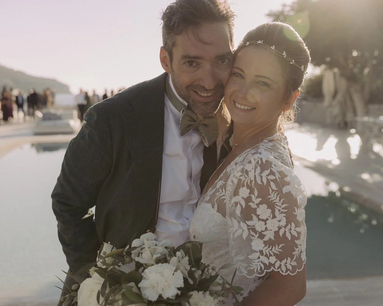 Happy couple at their wedding outdoors during sunset, with the man wearing a dark suit and bow tie, and the woman wearing a lace wedding dress and floral headband, holding a bouquet of white flowers.