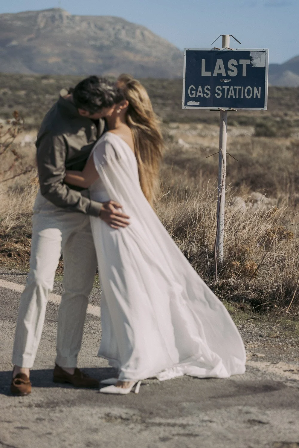 A couple kissing near a sign that reads 'Last Gas Station' in a desert landscape.