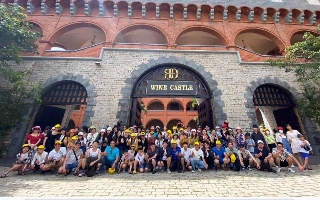 Group of people in front of Wertheimer's Wine Castle sign, posing for a group photo outside a large red brick building with arched entrances, surrounded by trees, on a cobblestone pavement.