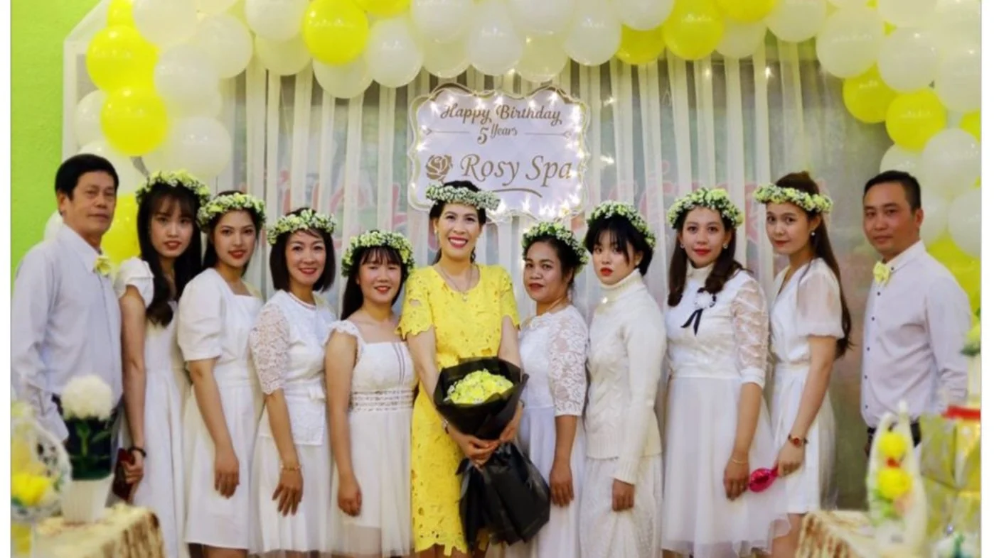 A group of people celebrating a birthday at a party with yellow and white decorations. They are standing in front of a banner that reads "Happy Birthday 5 Years Rosy Spa." The group includes women wearing white dresses with flower crowns, and a woman in a yellow dress holding a bouquet of flowers.
