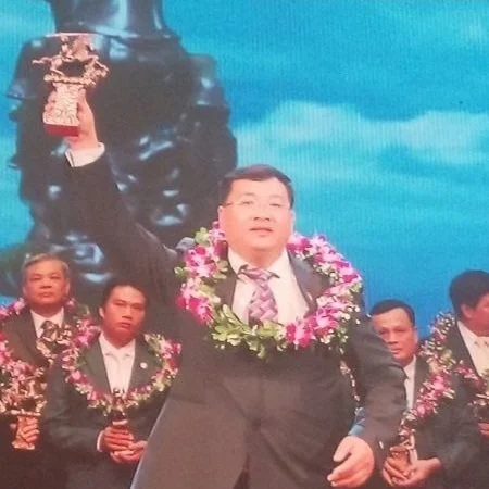 A man in a suit holding a trophy in one hand and wearing a floral garland around his neck on a stage with other similarly dressed men and a backdrop of a statue and sky.