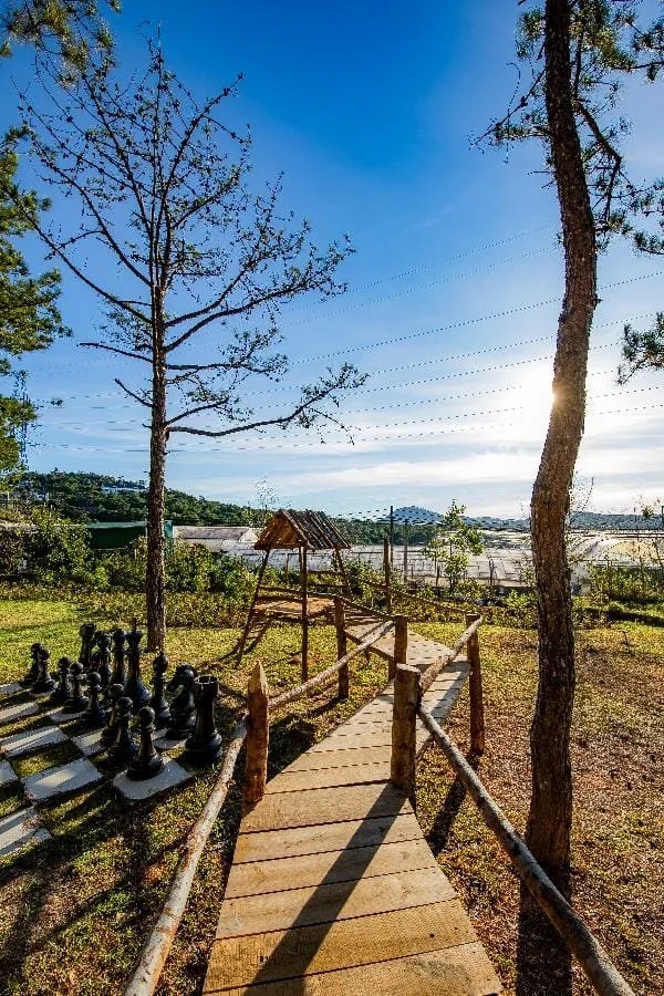 A small wooden bridge over grass, leading to a miniature children’s playground with a small wooden slide, surrounded by trees and a bright sunny sky.