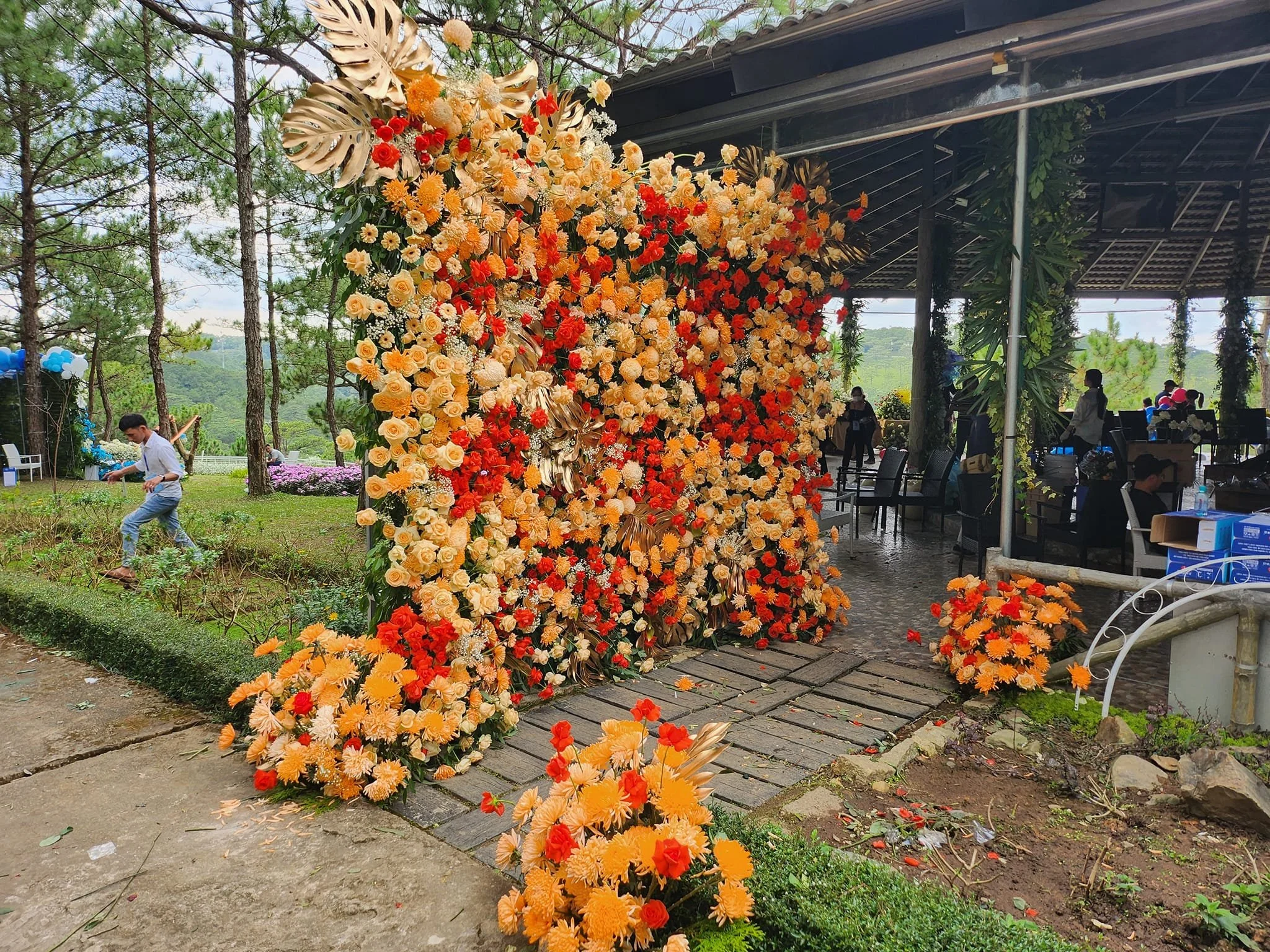 Colorful flower arch with orange, yellow, and red flowers at an outdoor event, with people and trees in the background.