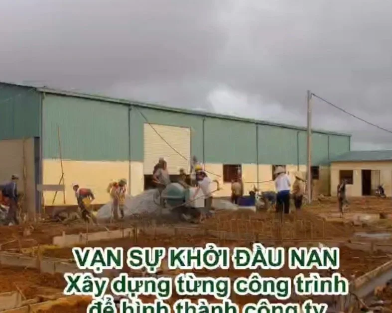 Construction site with workers pouring concrete in front of a large green and yellow warehouse building, under cloudy sky.