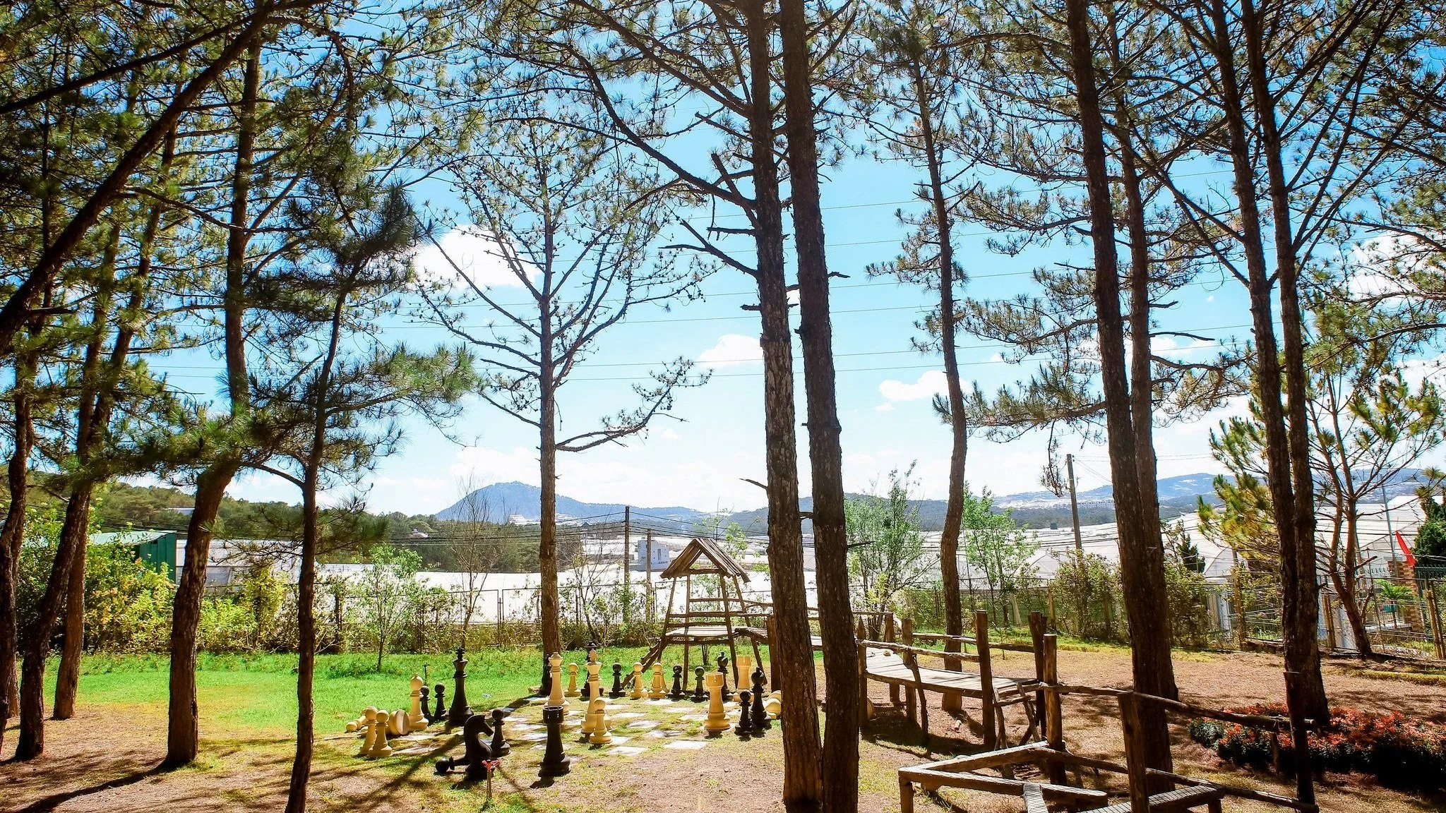 Outdoor scene with tall trees, a large chess set on a checkered ground, and mountains in the background under a blue sky with some clouds.