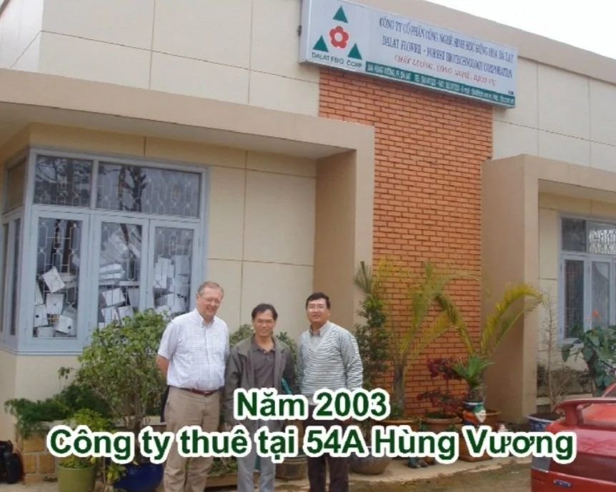 Photo of three men standing in front of a building with Vietnamese text at 54A Hung Vuong in 2003. The building has windows with papers in the panes and small plants and bushes outside. A sign with a logo and Vietnamese writing is visible above the men.