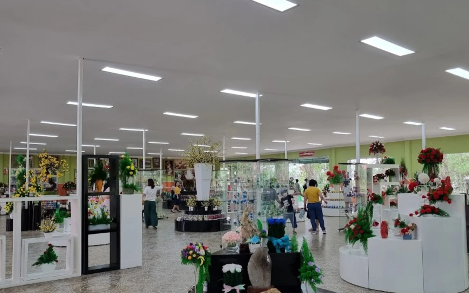 Interior of a gift shop decorated with various colorful artificial flowers and floral arrangements displayed on white shelves and stands, with customers browsing.