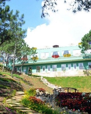 A multi-story light blue building with red, yellow, and green umbrellas on a balcony, surrounded by trees and a landscaped garden with stone steps and flowers.