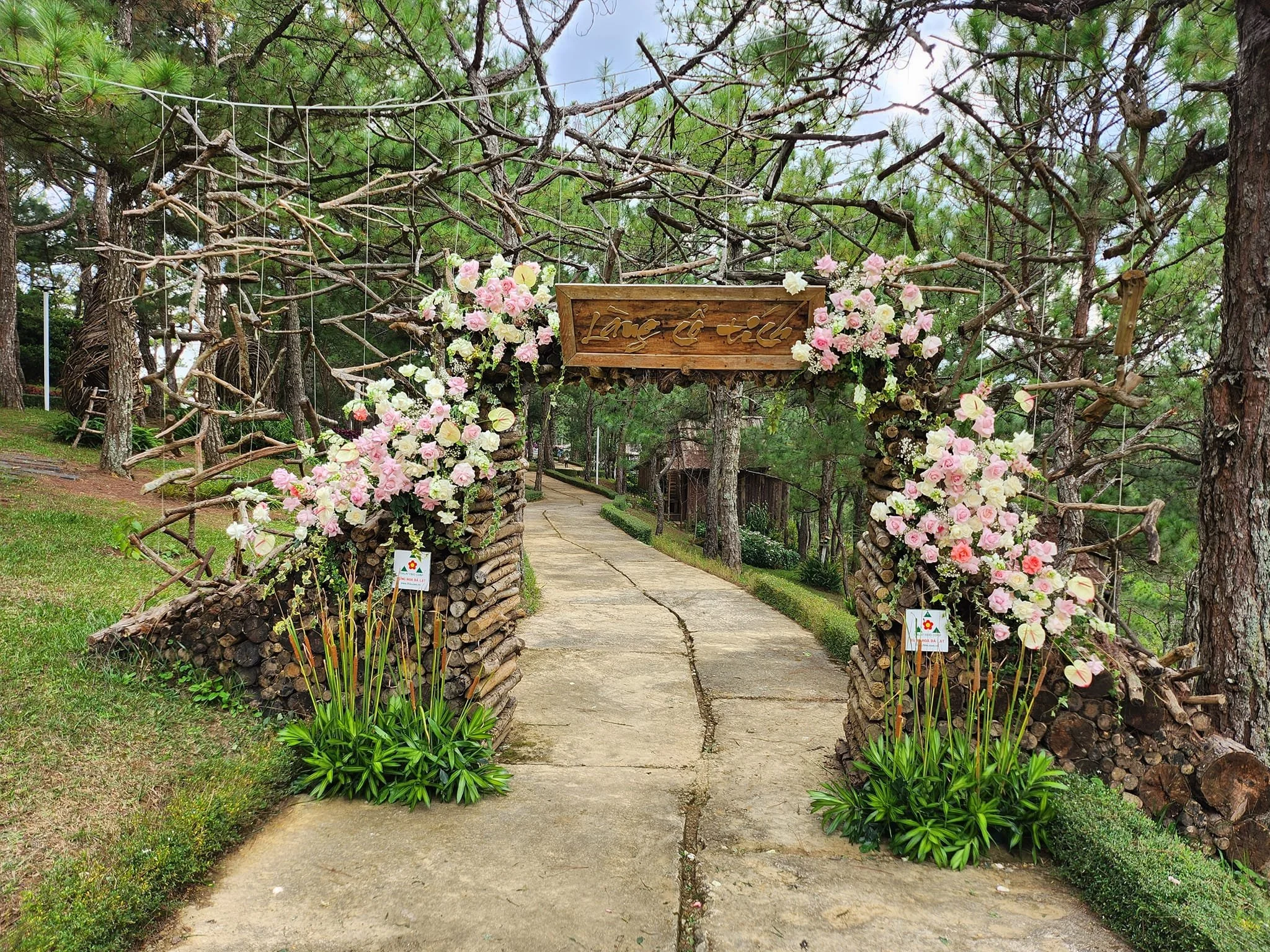 A pathway through a garden with a rustic wooden arch decorated with pink and white flowers and attached to logs, surrounded by pine trees.