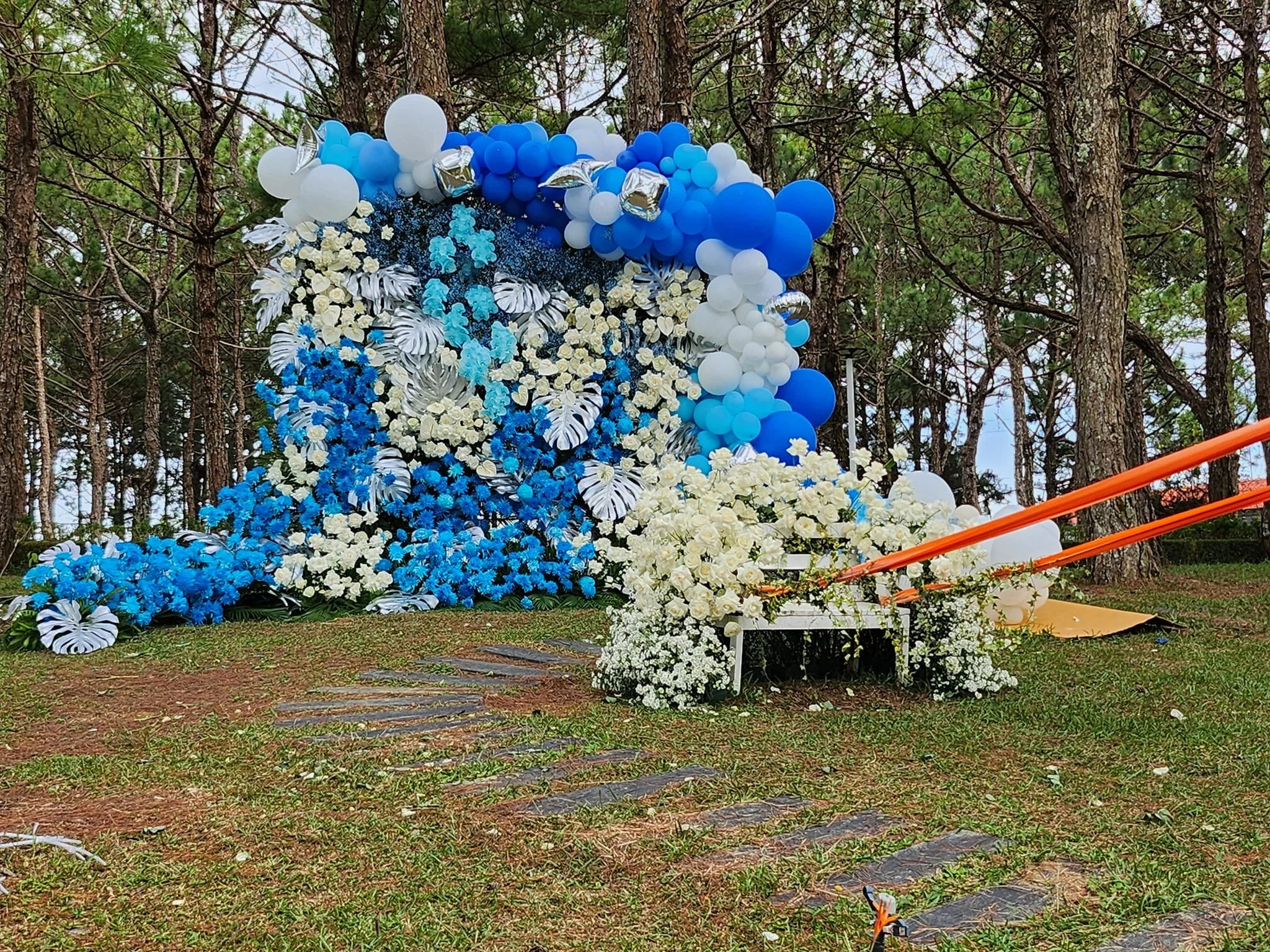 An outdoor decorative floral display with predominantly white and blue flowers, surrounded by white, blue, and silver balloons, set against trees in a forested area.