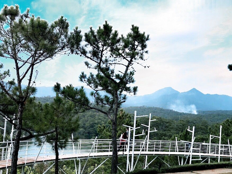 A scenic view of a white bridge surrounded by trees, with mountains and smoke in the background under a partly cloudy sky.