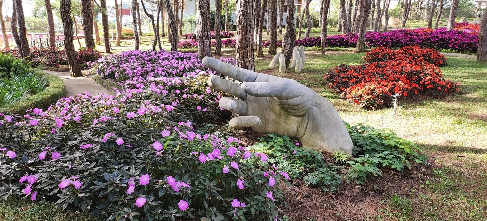A garden with colorful flowers including pink, red, and purple blooms, a large stone sculpture of a human hand making a peace sign, and a walking path surrounded by trees and bushes.