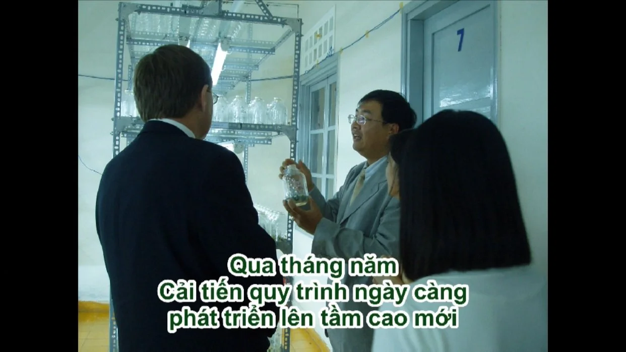 Three people in a laboratory discussing, with scientific glassware on metal shelves in the background. The person in the center is holding a bottle and speaking, while the others listen. Vietnamese subtitles are shown at the bottom.