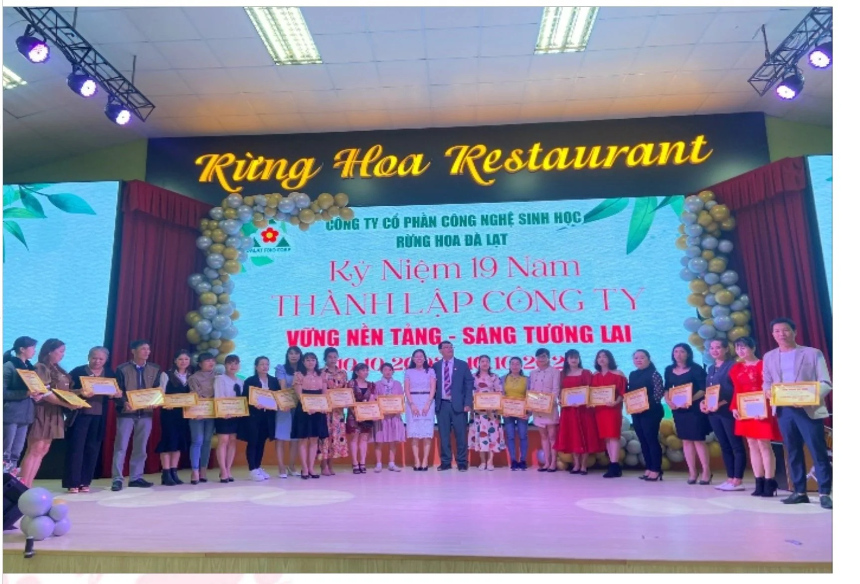 Group of people standing on stage holding certificates during a celebration event at Rừng Hoa Restaurant, commemorating the 19th anniversary of a company's founding, with decorations including balloon arrangements and a large LED screen.