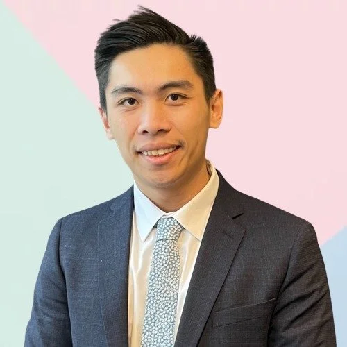 A portrait of a young Asian man in a dark suit, white shirt, and patterned tie, smiling against a colorful pastel background.