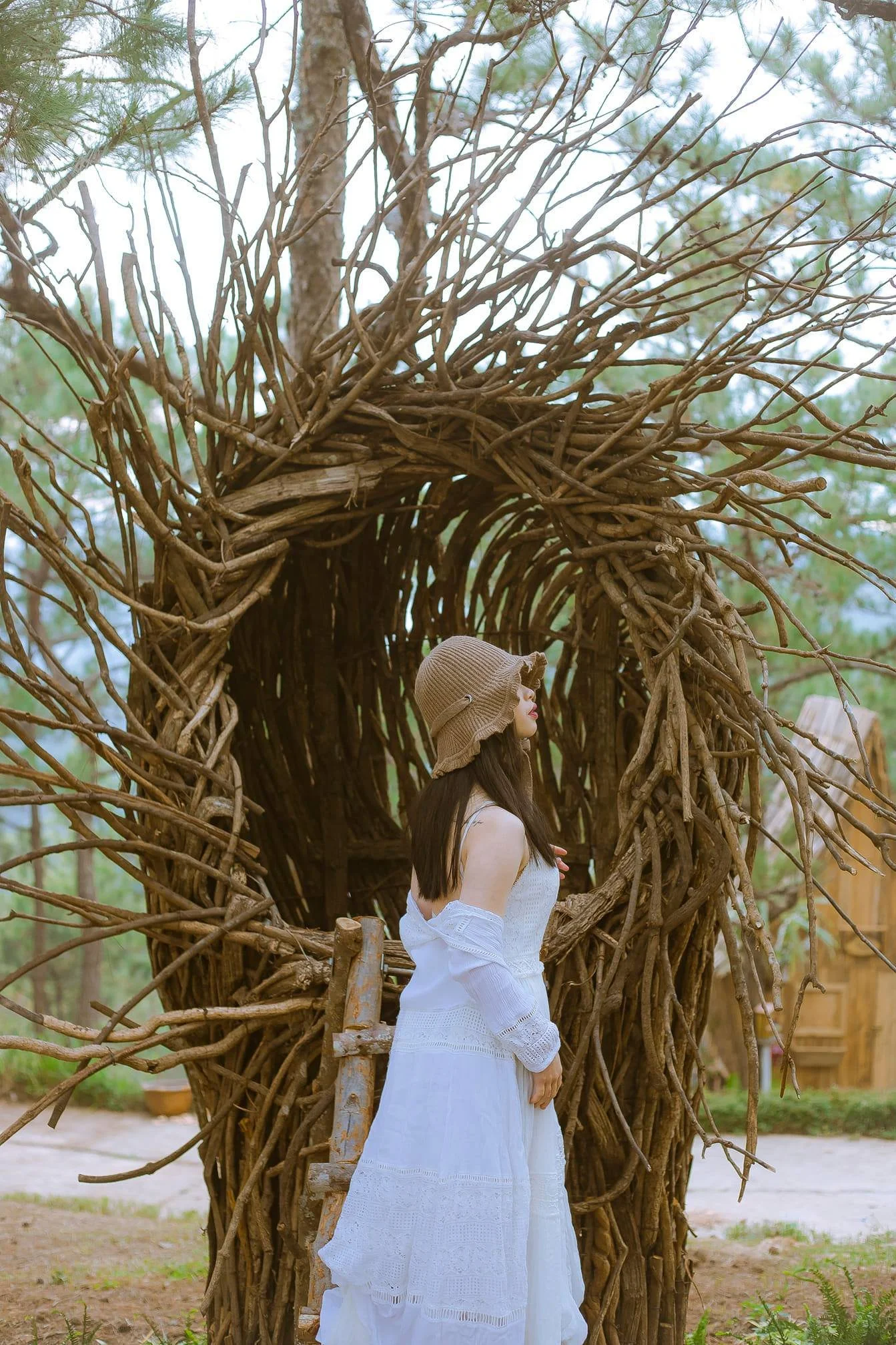 A woman in a white dress and a brown sun hat stands next to a large, woven, tree branch sculpture outdoors, with trees and a dirt ground in the background.