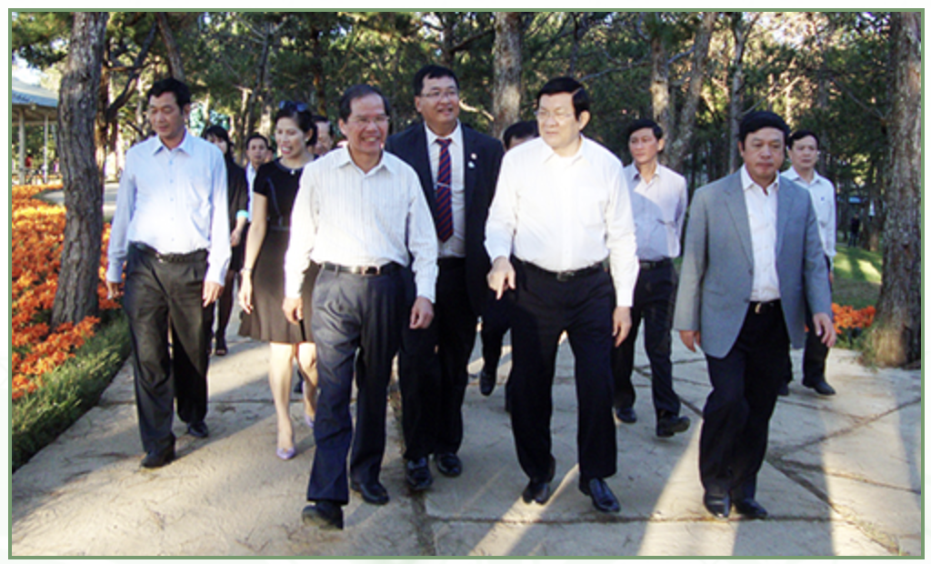 Group of professionally dressed people walking outdoors on a sunny day in a park or garden, smiling and engaging in conversation.